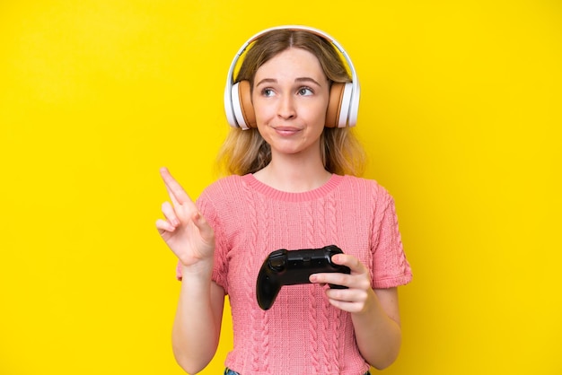 Blond Engels jong meisje speelt met een videogamecontroller geïsoleerd op gele achtergrond met gekruiste vingers en het beste wensen