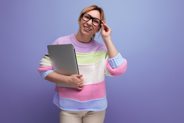 Blond female student with a laptop in her hands adjusts her glasses on a purple background