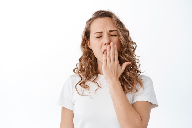 Blond girl feeling tired covering yawning mouth with hand feeling bored or exhausted fall asleep standing against white background