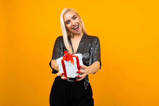 Blond girl holds out a gift box with a red ribbon on a yellow wall