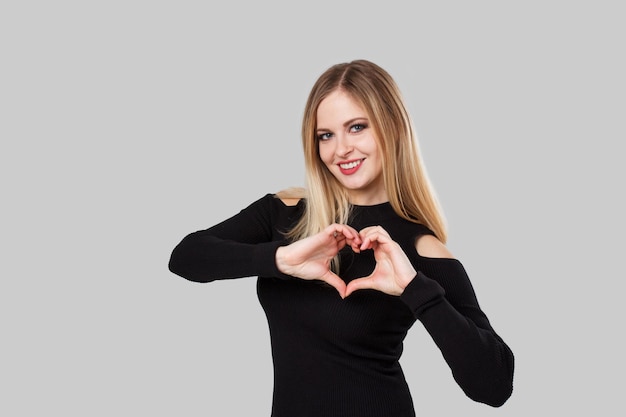 Blond girl is smiling and is making a heart sign with her hands. The background is light gray.