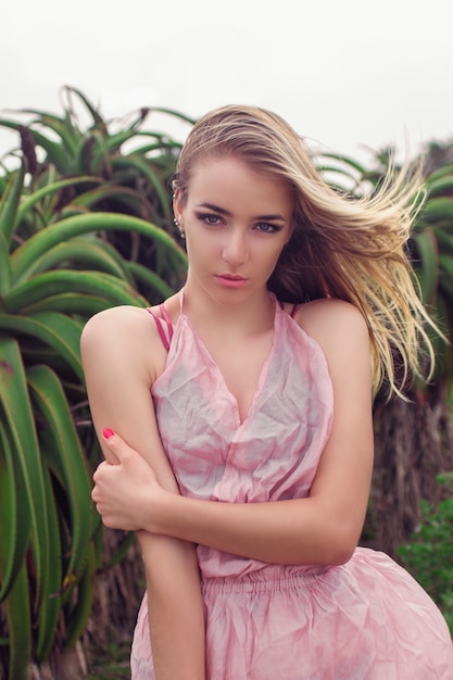 Blond girl in a light pink dress stands against the background of a green plant