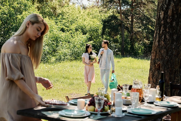 테이블에 집에서 만든 파이로 접시를 넣어 금발 소녀는 젊은 부부가 녹색 잔디 아래로 이동하는 동안 친구와 함께 저녁 식사를 위해 제공