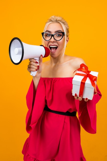 Blond girl in a red dress talks about gifts holding a megaphone and a gift box in hands on a yellow space