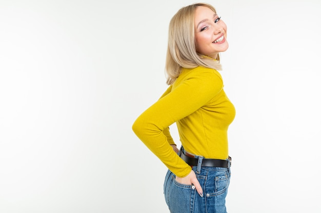 Blond girl in a tight-fitting yellow blouse and jeans poses sideways on a white background