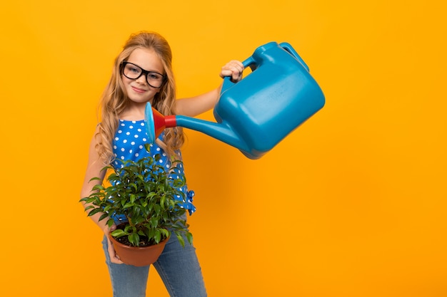 Blond girl watering a leaf plant from a watering can on an orange wall