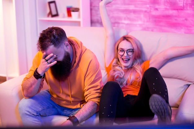 blond girlfriend happy to win against her boyfriend on video games sitting on sofa. room with neon lights.