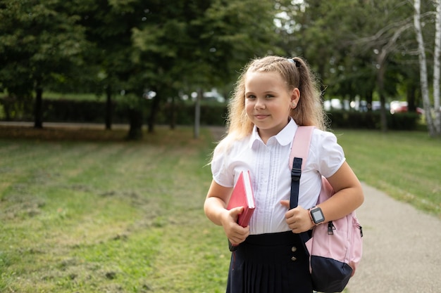 blond glimlachend schoolmeisje in schooluniform met notitieboekje met roze rugzak terug naar school