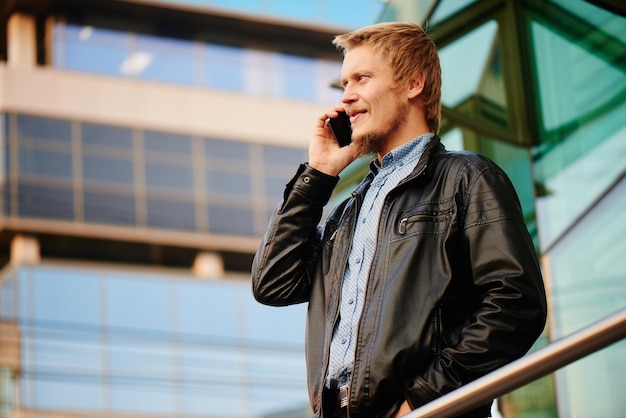 Блондин с бородой в кожаной куртке и рубашке разговаривает по телефону на заднем плане здания со стеклянным фасадом
