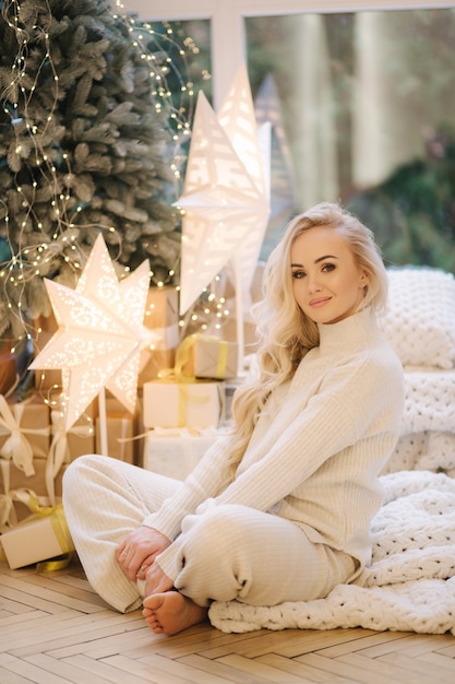 Blond haar vrouw in wit gebreid pak om thuis te zitten door kerstboom. Mooie vrouw in de winter