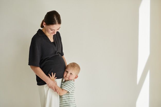 Blond-haired boy touching ear to belly of mother while listening to heartbeat of brother or sister, studio shot