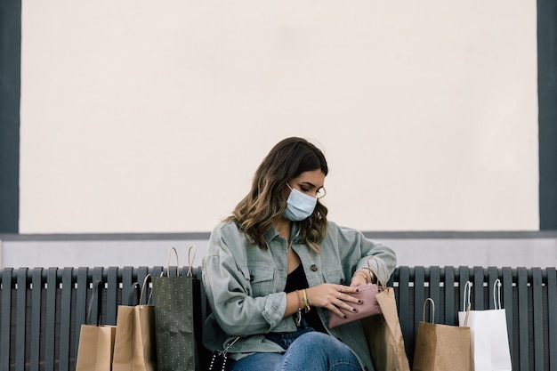 Blond-haired white woman with protective mask sitting on a bench outdoors surrounded by shopping paper bags looking at her purse.