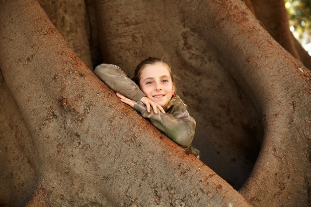 Photo blond kid girl smiling arms on big ficus tree