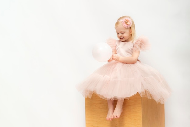 Blond kind in weelderige jurk zit op houten kubus geïsoleerd op witte achtergrond Meisje kijkt naar ballon in handen Kopieer de ruimte