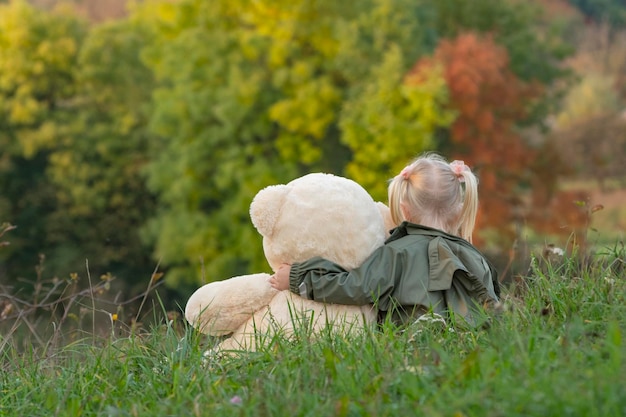 Blond klein meisje en teddybeer op gras op herfstbomen achtergrondkleur Kind met zacht stuk speelgoed rust in de natuur
