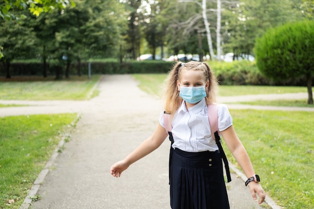 Blond krullend schoolmeisje met beschermend masker in schooluniform met roze rugzak terug naar school