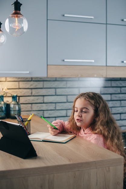 Blond lief meisje gebruikt een tablet tijdens online lessen in de keuken vanuit huis from