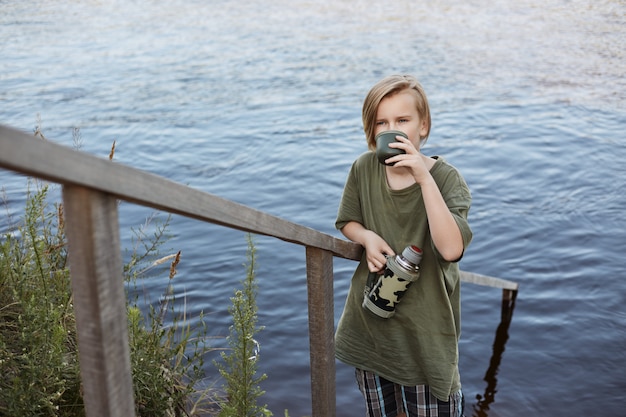 Blond little boy drinking hot tea from thermos isolated over river, male child spending time in open air, wearing green t shirt, enjoying hot beverage while posing near water.