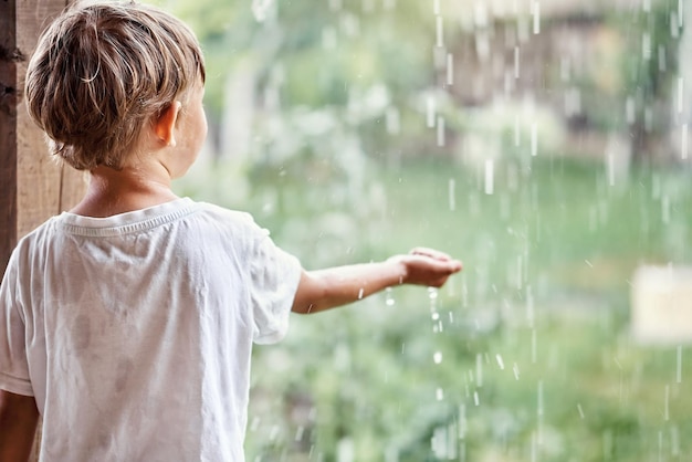 Blond little boy holds hand under falling down rain drops standing on country house terrace