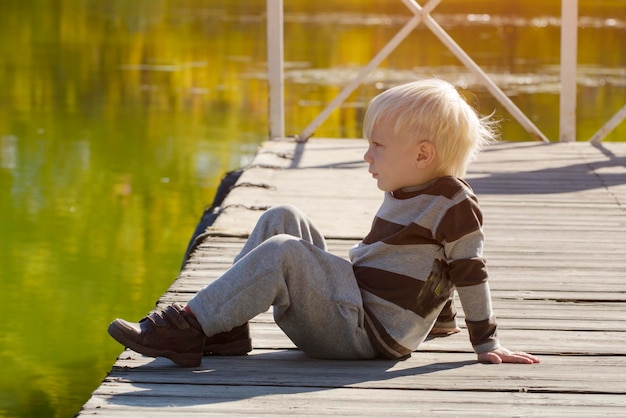 Белокурый маленький мальчик сидит на берегу реки Осенний солнечный день на пирсе