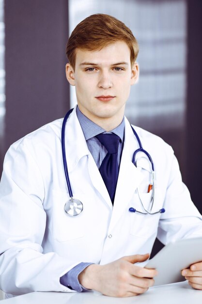 Blond male doctor sitting and working with tablet computer in clinic at his working place, close-up. Young physician at work. Perfect medical service, medicine concept.