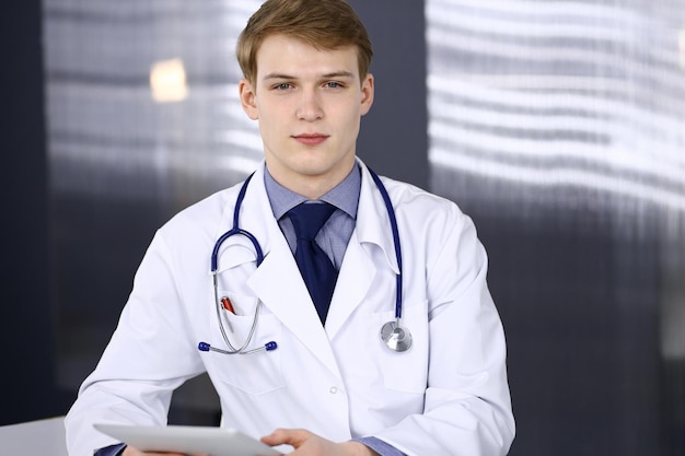 Blond male doctor sitting and working with tablet computer in clinic at his working place, close-up. Young physician at work. Perfect medical service, medicine concept.