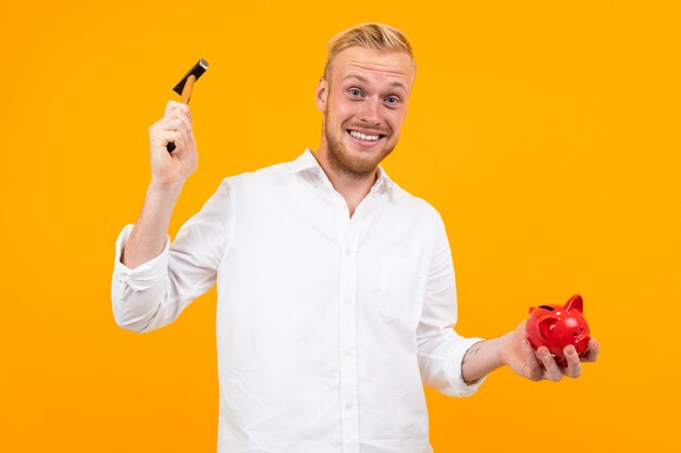 Blond man in a white shirt smashes a piggy bank with a hammer on yellow