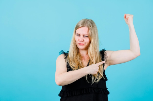 Foto blond meisje heft haar spier op en toont het met wijsvinger op blauwe achtergrond