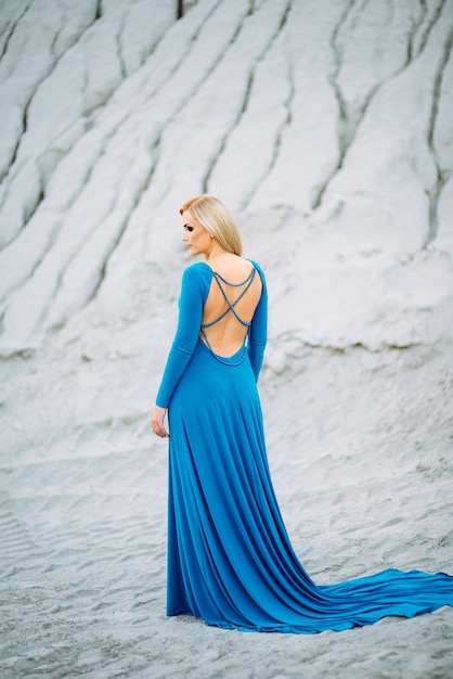 Blond meisje in een blauwe jurk met blauwe ogen in een granietgroeve tegen de achtergrond van grind