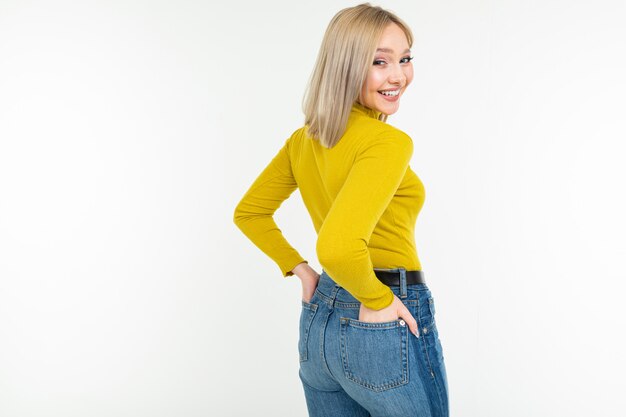 Blond meisje in een nauwsluitend geel jasje en jeans die zijdelings op een witte achtergrond stellen