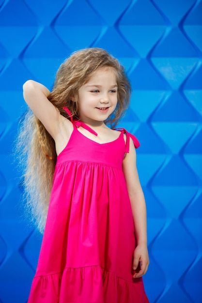 Blond meisje in een roze jurk op een blauwe achtergrond dansen en lachen, heldere kinderemoties van vreugde, gelukkige jeugd