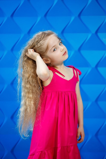 Blond meisje in een roze jurk op een blauwe achtergrond dansen en lachen, heldere kinderemoties van vreugde, gelukkige jeugd