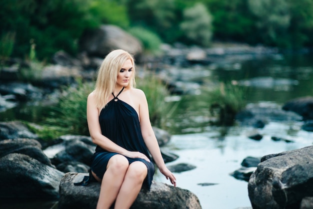 Blond meisje in een zwarte jurk met blauwe ogen aan de stenen kust van de rivier