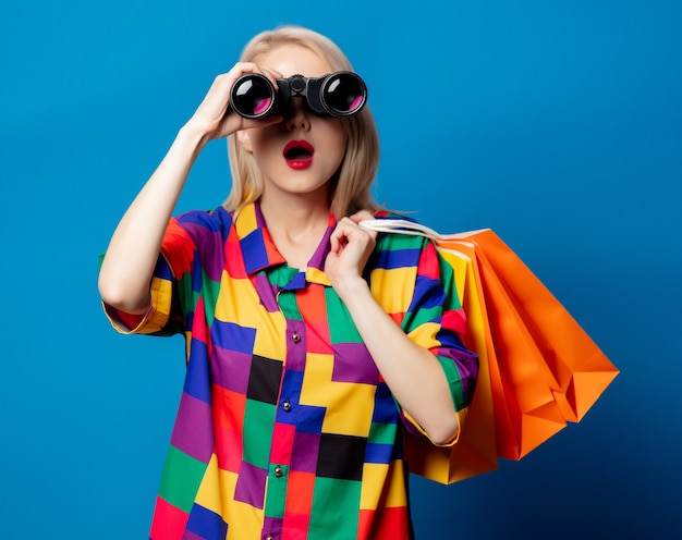 Blond meisje in jaren 90 shirt met verrekijker en boodschappentassen op blauwe ruimte