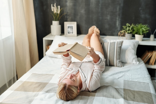 Blond meisje in pyjama liggend op bed en boek lezen tijdens de voorbereiding op examen of plezier