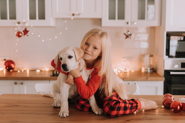 Blond meisje in rode kerstpyjama zit in een keuken met bloemenkrans met een witte hond. winterweekenden en vakanties in een warme familiekring. kind en huisdier. ruimte voor tekst. Hoge kwaliteit foto