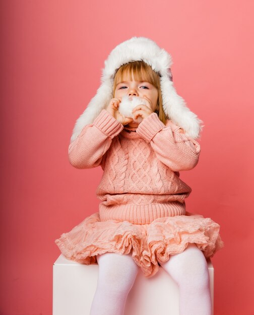 Blond meisje in winterkleren en een bontmuts op een roze achtergrond.