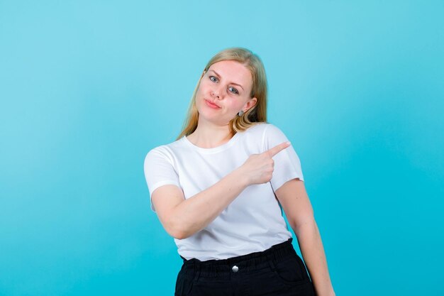 Blond meisje kijkt naar de camera door met de wijsvinger naar rechts te wijzen op een blauwe achtergrond