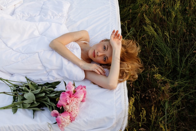 Blond meisje ligt op het bed met wit linnen op het groene gras. bij het meisje een boeket roze bloemen.