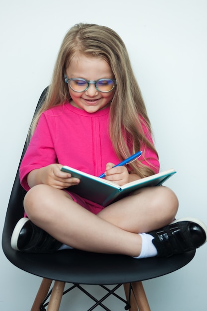 Blond meisje met bril schrijft iets in een boek terwijl ze poseren in een fauteuil op een witte muur