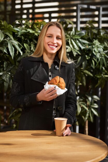 Blond meisje met een papieren kopje koffie en muffin zittend in een café. straatvoedselconcept