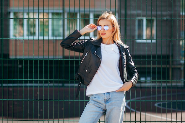 Blond meisje met t-shirt, bril en leren jas poseren tegen straat