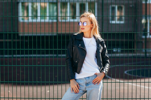 Blond meisje met t-shirt, bril en leren jas poseren tegen straat