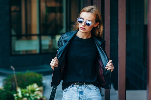 Blond meisje met zwarte t-shirt, bril en lederen jas poseren tegen stree