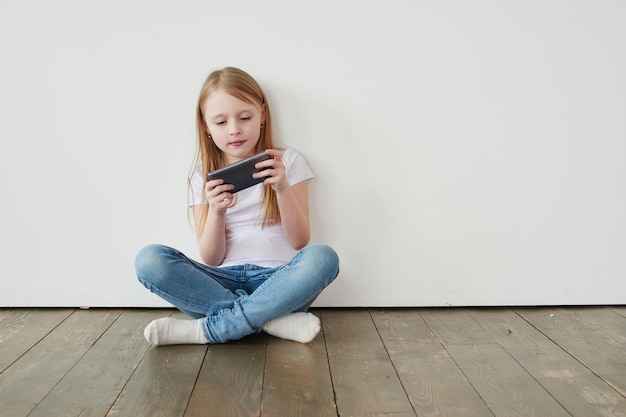 Blond meisje spelen telefoon zittend op houten vloer op witte achtergrond