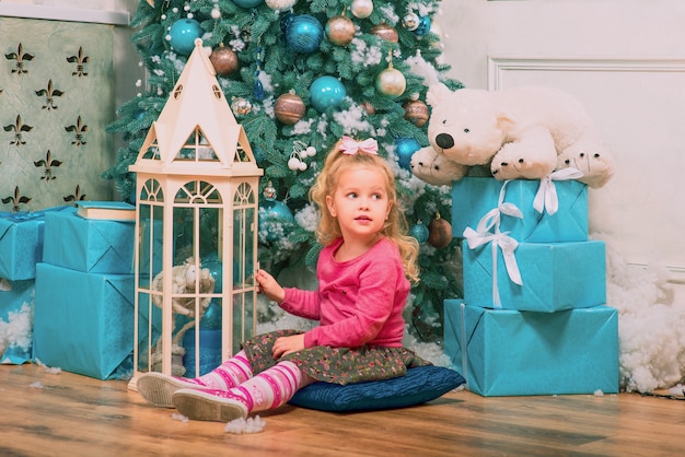 Blond meisje zit en lacht bijna versierde kerstboom en nieuwjaarscadeautjes