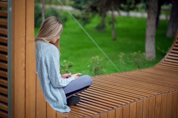 Blond meisje, zittend met benen op een bankje en kijken naar een smartphone