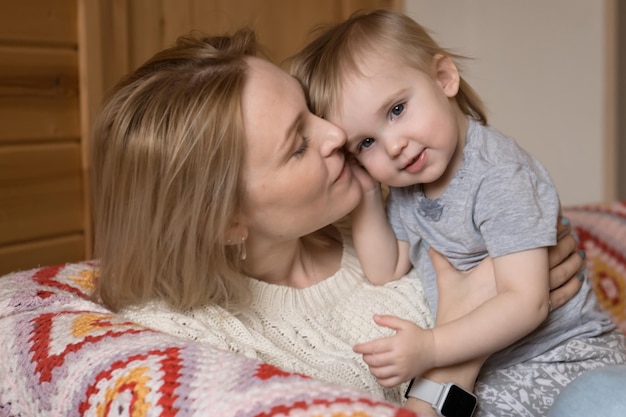 Blond mother kissing little daughter or son sitting on sofa at home