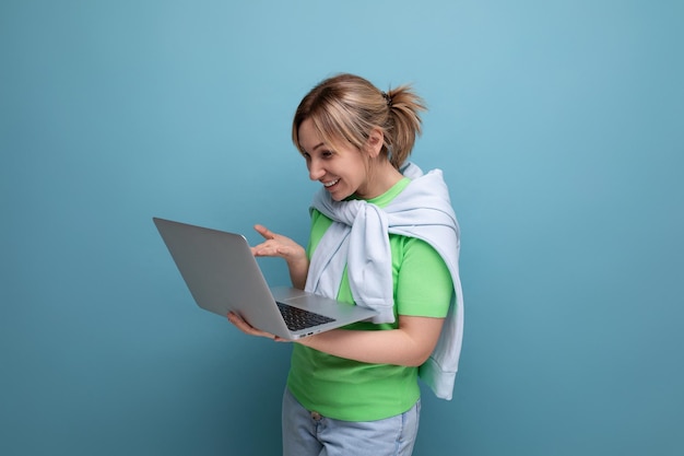 Blond pensive girl in casual outfit uses app on laptop on blue background