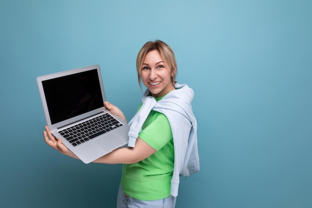 캐주얼 의상을 입은 금발의 긍정적인 매력적인 소녀는 파란색 노트북에 흉내낸 화면을 보여줍니다.